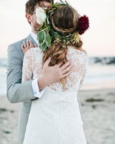 a bride and groom hugging on the beach with flowers in their hair, dressed in a gray suit