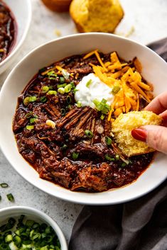 a bowl of chili with cornbreads and sour cream on the side is being held up by someone's hand