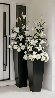 two tall black vases with white flowers in them sitting on the ground next to a door