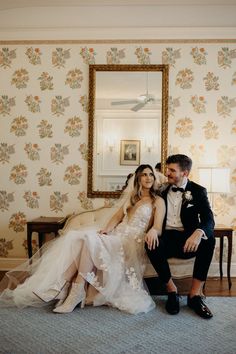 a bride and groom sitting on a couch in front of a wallpapered room