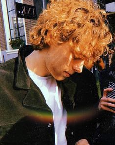 a man with curly red hair looking at his cell phone while standing in front of a building