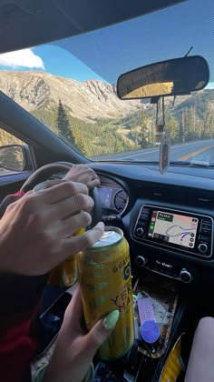 a person driving a car with two cans of sodas in the passenger seat and mountains in the background