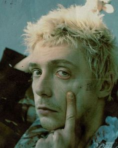 a young man with blonde hair and blue eyes looks at the camera while holding his hand to his face