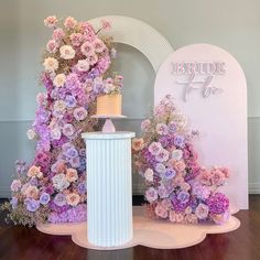 a cake on top of a pedestal surrounded by pink and purple flowers in front of a bride to be sign