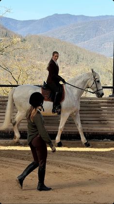 two women are riding on horses in an outdoor arena with mountains in the back ground