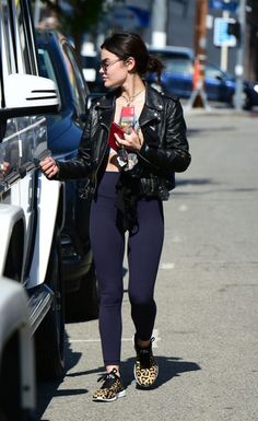 a woman in black jacket and leggings walking down street next to parked cars