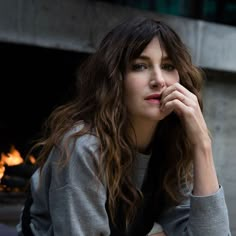 a woman with long hair sitting in front of a fire place and looking at the camera