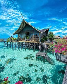 a house on stilts in the water with pink flowers and blue sky behind it