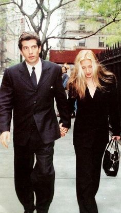 a man and woman are walking down the street together holding hands while wearing black suits