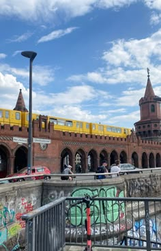 people are walking on the sidewalk near a wall with graffiti and a yellow train in the background