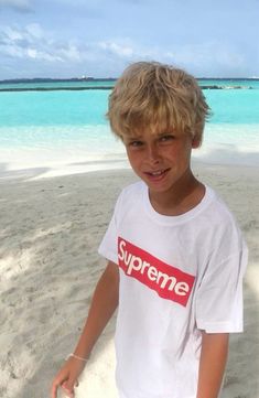 a young boy standing on top of a sandy beach next to the ocean with blue water in the background