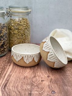 two ceramic bowls sitting on top of a wooden table next to jars filled with seeds