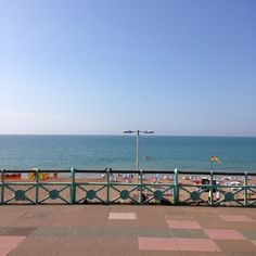 people are sitting on benches near the beach and looking out at the ocean from a pier