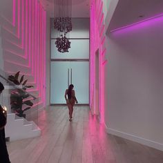 a woman walking down a hallway with pink lighting on the walls and stairs in front of her