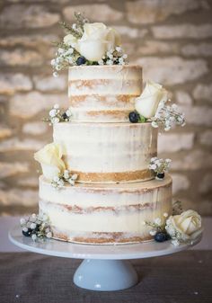 a three tiered cake with white flowers and blue berries on the top is displayed in front of a brick wall