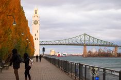 two people walking down a sidewalk next to the water with a bridge in the background