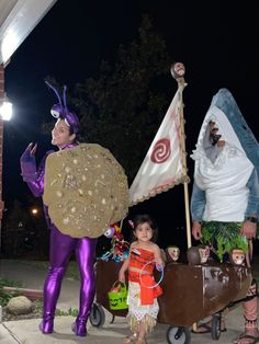 two people in costumes standing next to each other on a sidewalk at night with one holding a wheelbarrow and the other carrying a suitcase