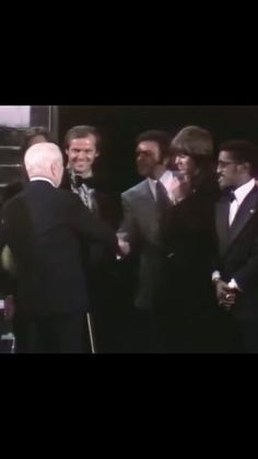 two men shaking hands in front of other men and one man wearing a tuxedo