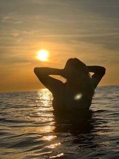 a woman is sitting in the water with her hands behind her head as the sun sets