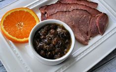 a white plate topped with meat, beans and an orange next to another plate filled with food