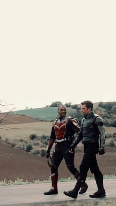 two men in black and red outfits are walking down the street with one man wearing boots