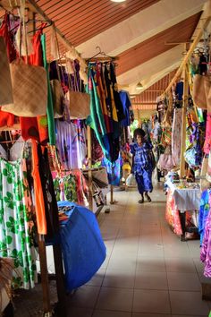 a woman walking through a market filled with lots of colorful clothing hanging from the ceiling