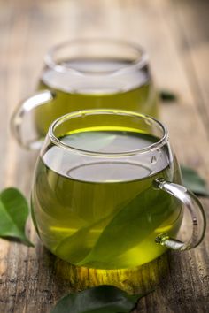 two glass mugs filled with green tea sit on a wooden table next to leaves