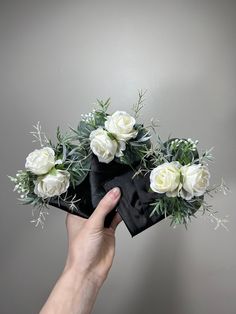 a hand is holding a black bow with white roses on it and greenery in the center