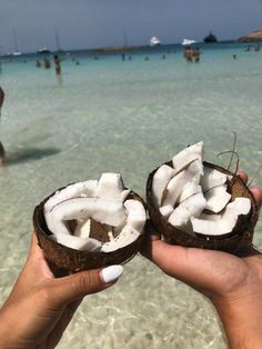 two people holding coconuts in their hands on the beach with other people swimming in the water behind them