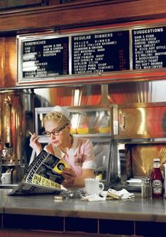 a woman is sitting at a counter in front of a menu board and pouring something into a cup