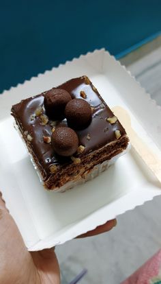 a person holding a square piece of cake with chocolate frosting and nuts on top