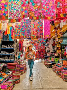 a woman walking through a market filled with lots of colorful hats and baskets hanging from the ceiling