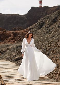 a woman in a long white dress walking across a wooden bridge with a lighthouse in the background