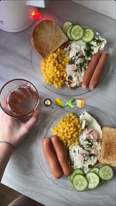 two plates with hotdogs, corn and vegetables on them next to a glass of water