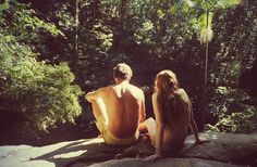 two people sitting on top of a rock near the water and trees in the background