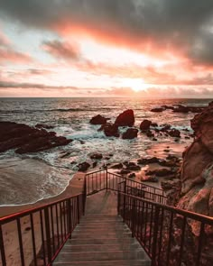 the stairs lead down to the beach as the sun sets over the water and rocks