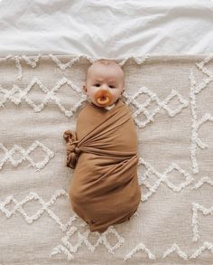 a baby wrapped in a brown wrap laying on top of a white blanket with a pacifier in it's mouth