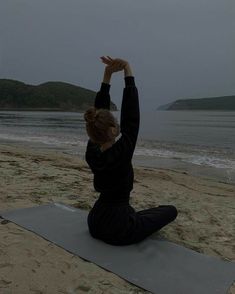 a woman sitting on top of a yoga mat in front of the ocean with her hands up