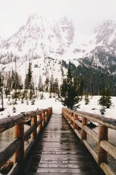 a wooden bridge with snow covered mountains in the backgrouds and trees on either side