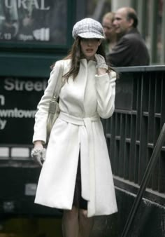 a woman in white coat and hat walking down the street with her hand on her hip