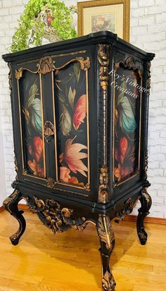 an ornate black and gold painted cabinet with flowers on the front, sitting on a hard wood floor next to a brick wall