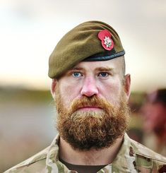 a bearded man with a red poppy on his cap looks at the camera while wearing an army uniform