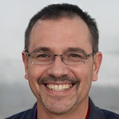 a man with glasses and a mustache smiling at the camera while standing in front of a white wall