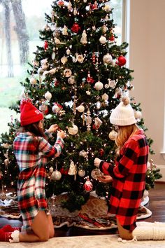 two women decorating a christmas tree in front of a large window with red and white ornaments