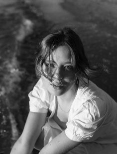 a woman in white shirt sitting on beach next to water and looking at the camera