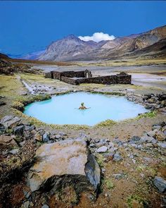 a man swimming in a blue pool surrounded by rocks and grass with mountains in the background