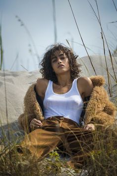 a woman sitting in the sand with her teddy bear wearing a white tank top and brown pants