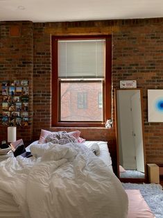 an unmade bed in front of a brick wall and window with pictures on it