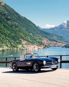 an old black sports car is parked by the water