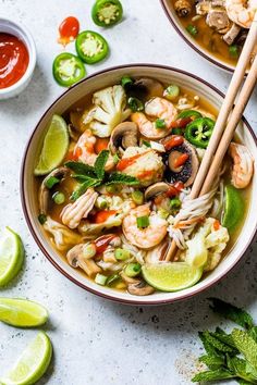 two bowls filled with shrimp and noodle soup next to limes, peppers, and sauce